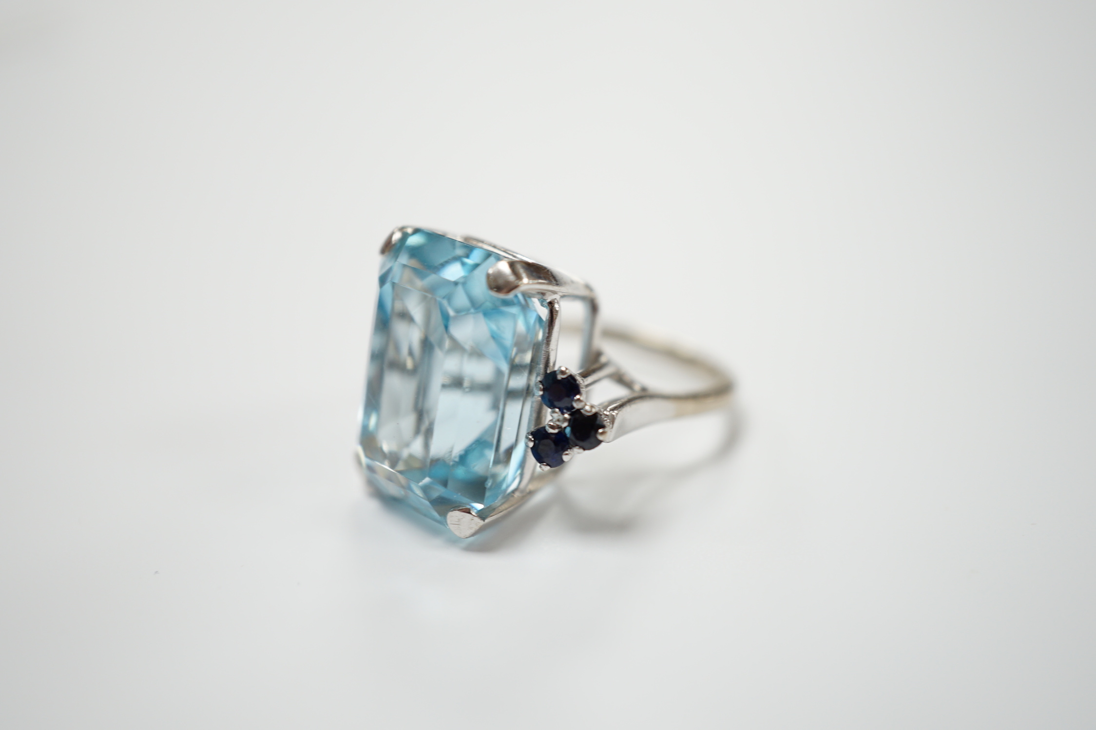An 18ct white metal and single stone emerald cut aquamarine set dress ring, with six stone sapphire set shoulders, size L, gross weight 11.8 grams.